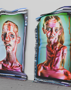 Two large, distorted digital portraits of humanoid figures printed on cloth-like material, leaning against a white wall on a concrete floor. The portraits feature exaggerated facial features and elongated necks with a glitch art aesthetic, including visible interface elements from digital editing software around the edges. The left figure has short hair and a tense expression, while the right figure has long hair and a more neutral expression. Both images have vibrant, surreal colours and warped textures.
